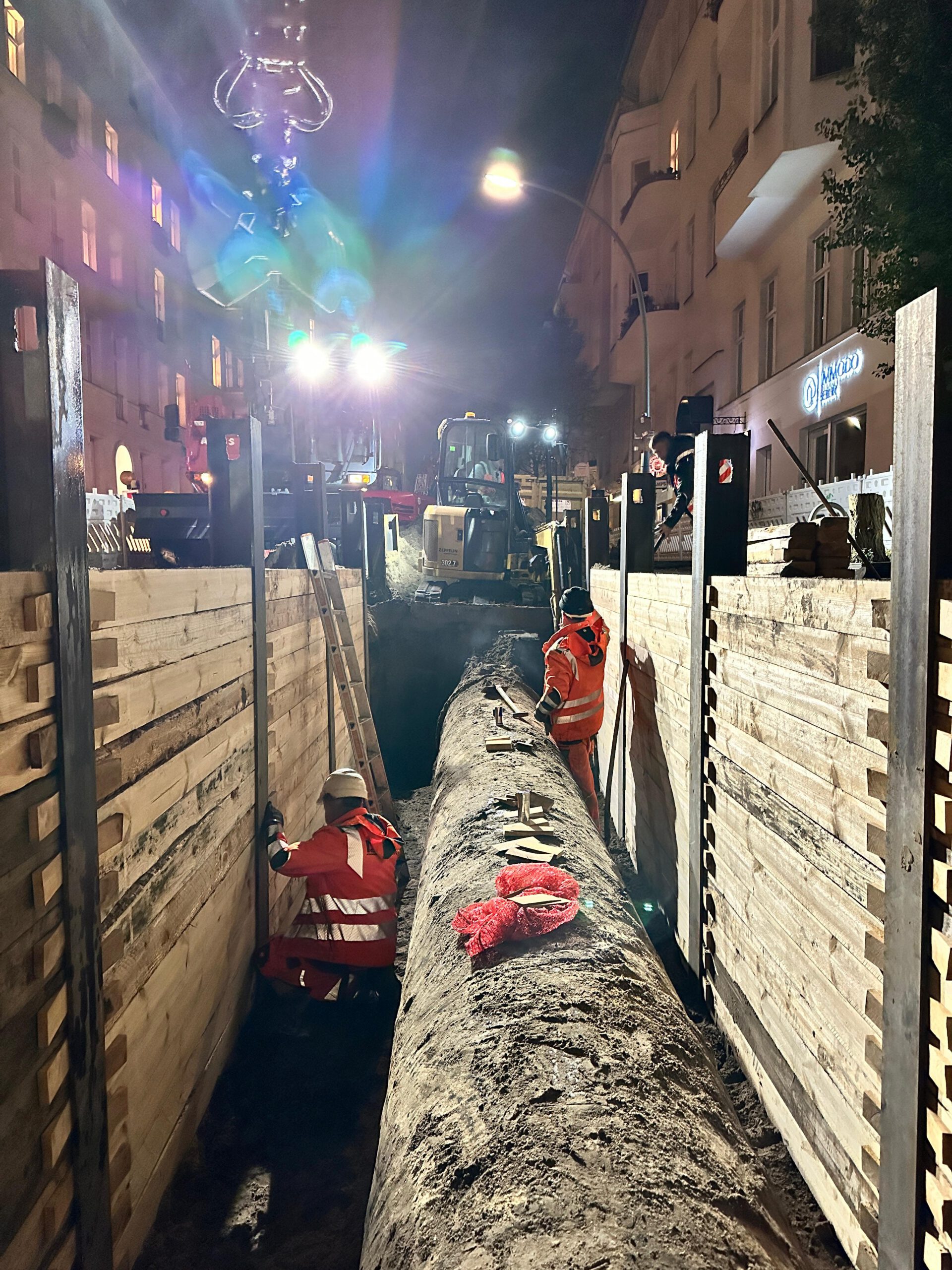 Nächtliche Rohrleitungsarbeiten mit Bagger und Team im gesicherten Graben